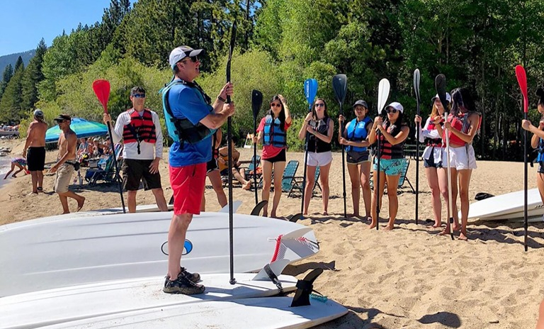 An instructor teaches and demonstrates to a group on a sandy beach the proper safety and use of stand up paddle boards.