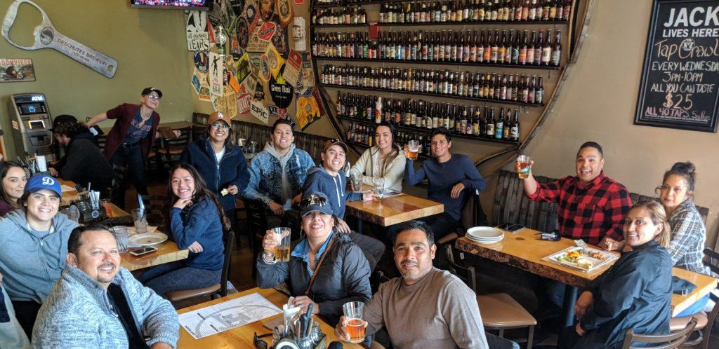 15 passengers enjoying beers and appetizers raising their glasses as they sit indoor tables.