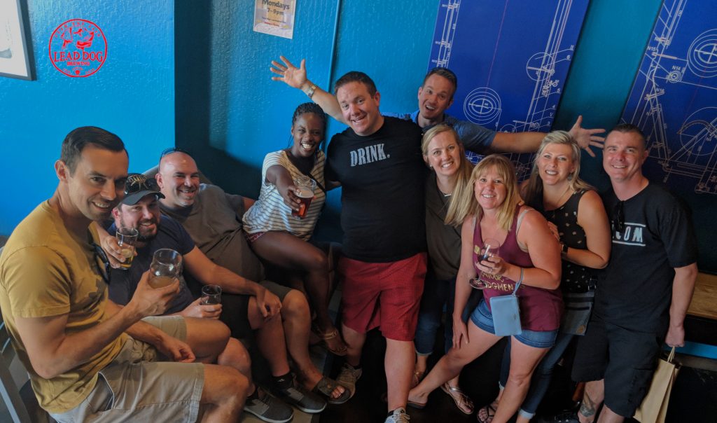 A group of 10 posing with beers raised, up against a blue wall inside the brewery tasting room.